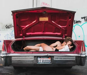 Man and woman embracing and kissing inside the open trunk of a red vintage car against a concrete wall with graffiti.