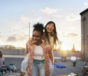 Two women laughing as one gives the other a piggyback ride on a sunny rooftop terrace at golden hour, photographed by Johnny Valentine.