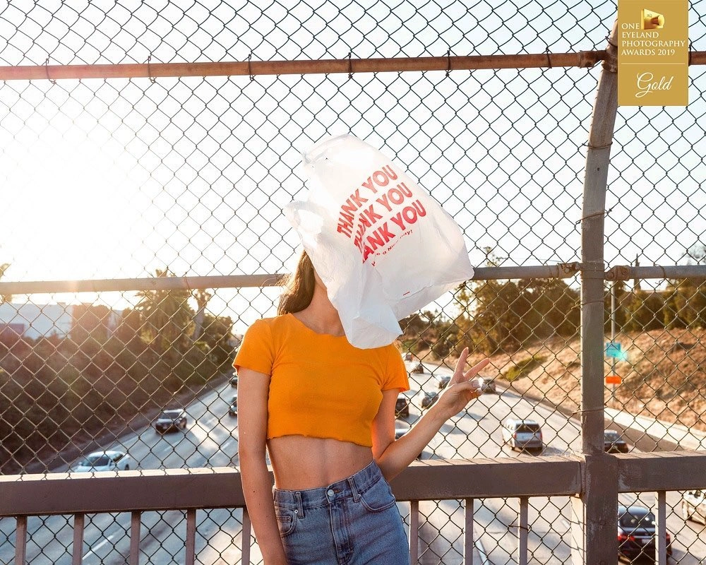 Woman in an orange crop top holding a white plastic bag over her face with a highway and chain link fence in the background