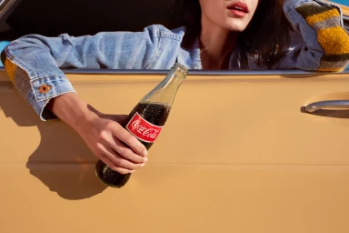 Woman in denim jacket leaning out of a yellow car window holding a glass Coca Cola bottle