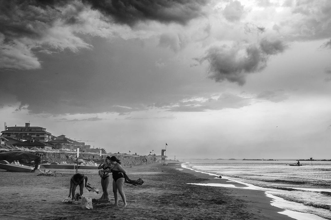 Black and white beach scene with three women in bathing suits, sand, sea, cloudy sky, and distant buildings creating a vintage feel.