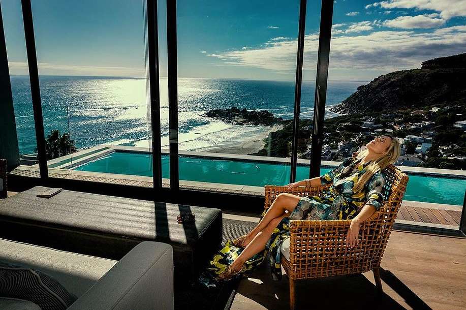 Adult woman in floral dress relaxes in a wicker chair indoors with a view of the ocean and swimming pool through large windows.