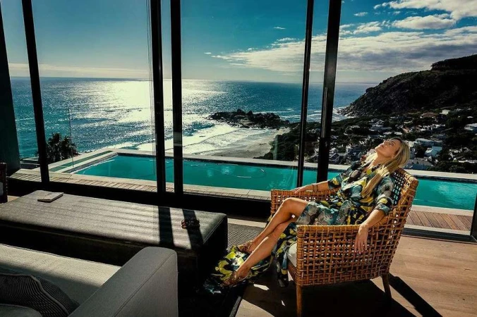 Adult woman in floral dress relaxes in a wicker chair indoors with a view of the ocean and swimming pool through large windows.
