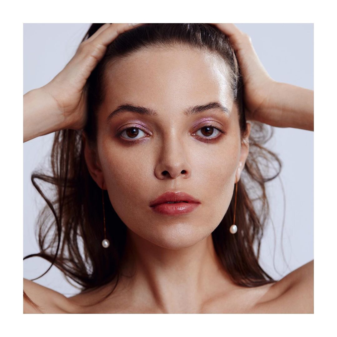 Close-up studio shot of a woman with brown hair holding her hands near her head, wearing pearl earrings and pink eyeshadow.