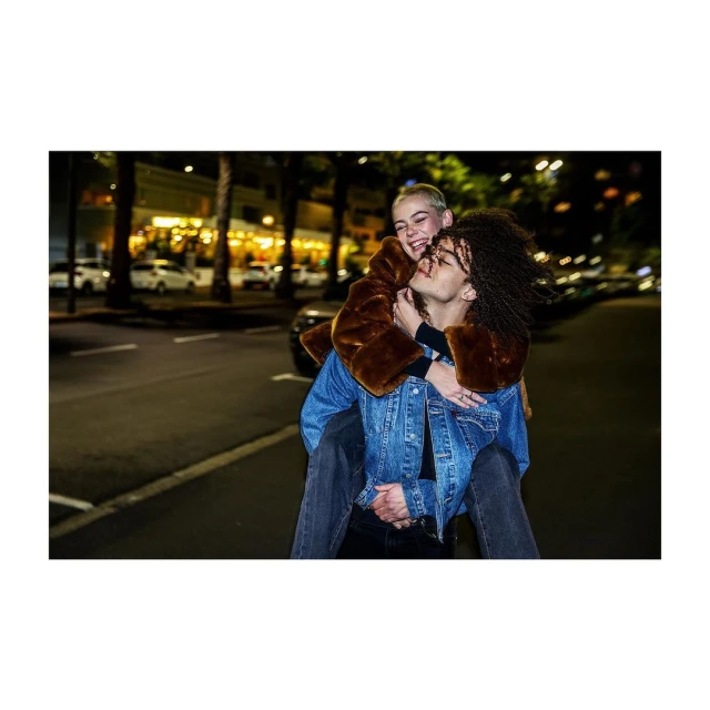 Two young adults share a playful moment on a city street at night, one giving the other a piggyback ride. Streetlights illuminate the scene.