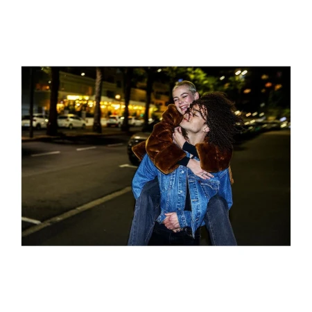Two young adults share a playful moment on a city street at night, one giving the other a piggyback ride. Streetlights illuminate the scene.