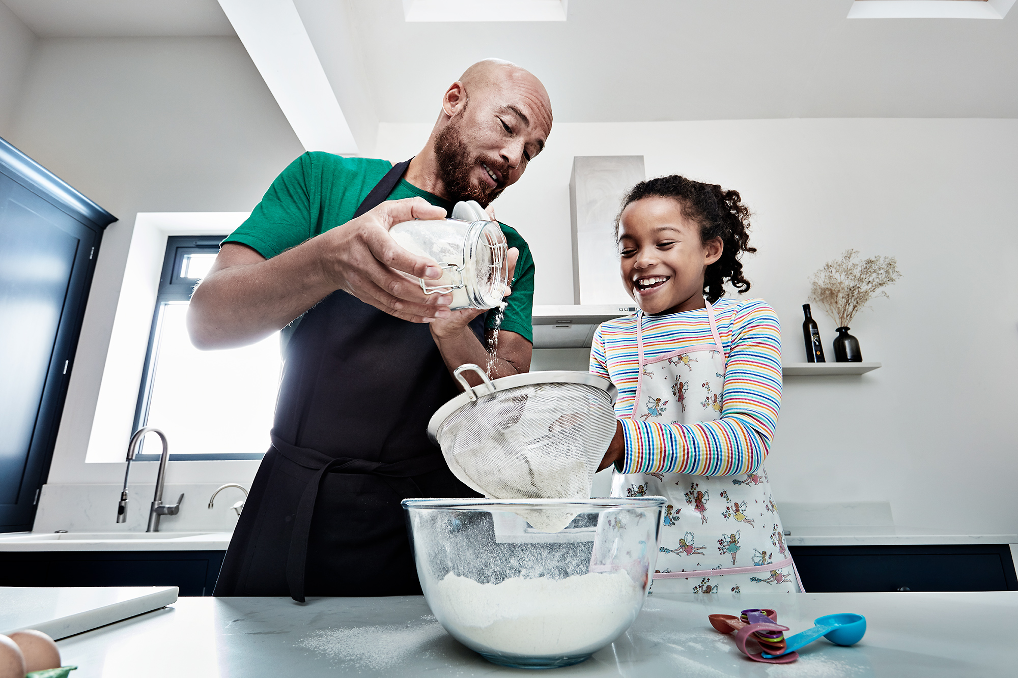 A father and daughter smile while baking in a bright kitchen, the father pouring ingredients through a sieve into a bowl on the countertop.