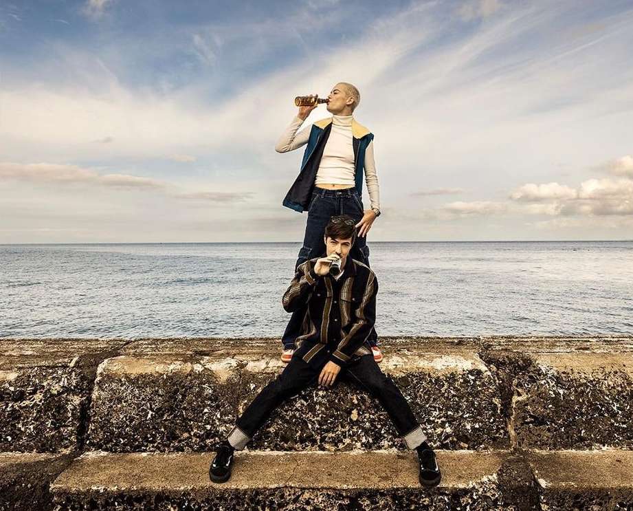 Two people, one on the other's shoulders, drink from bottles outdoors with the ocean and cloudy sky in the background, medium shot.