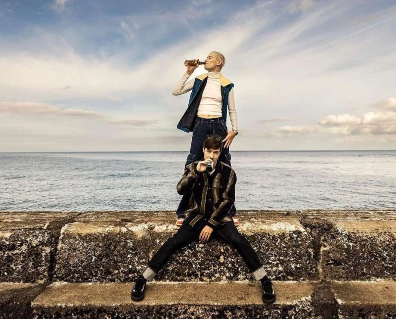Two people, one on the other's shoulders, drink from bottles outdoors with the ocean and cloudy sky in the background, medium shot.