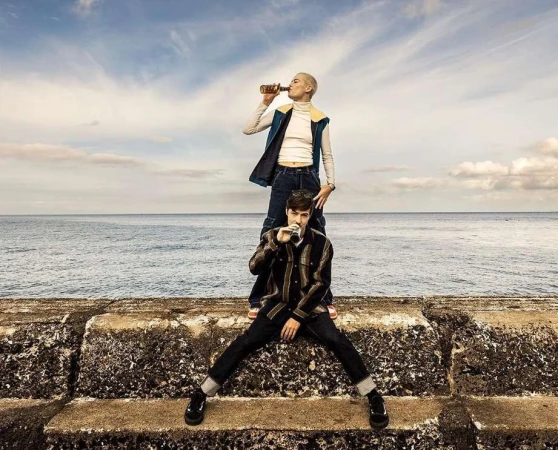Two people, one on the other's shoulders, drink from bottles outdoors with the ocean and cloudy sky in the background, medium shot.