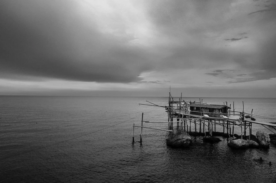 Black and white photograph of a trabucco fishing platform extending into the sea under an overcast sky. Landscape orientation.