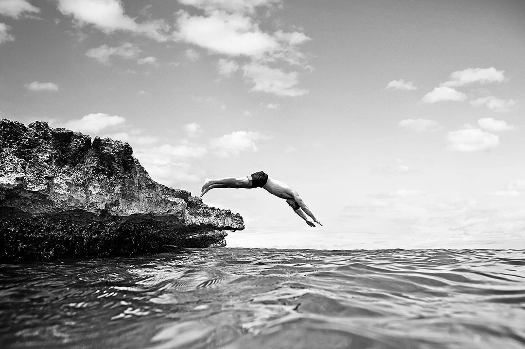 Black and white photograph of an adult male diving from a rocky cliff into rippled water under a cloudy sky in an outdoor setting.