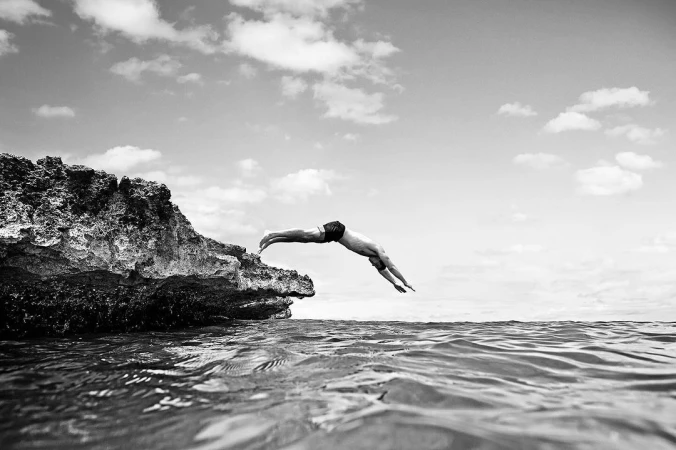 Black and white photograph of an adult male diving from a rocky cliff into rippled water under a cloudy sky in an outdoor setting.