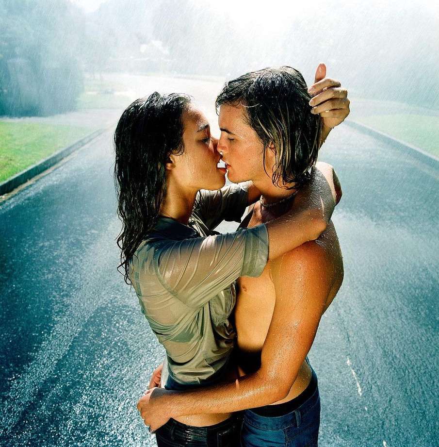 Two young adults embracing and kissing passionately in the rain on a paved road, creating an intimate, romantic outdoor scene.