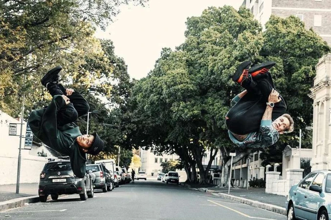 Two men perform parkour stunts in an urban environment, flipping upside down on a city street lined with parked cars and trees.