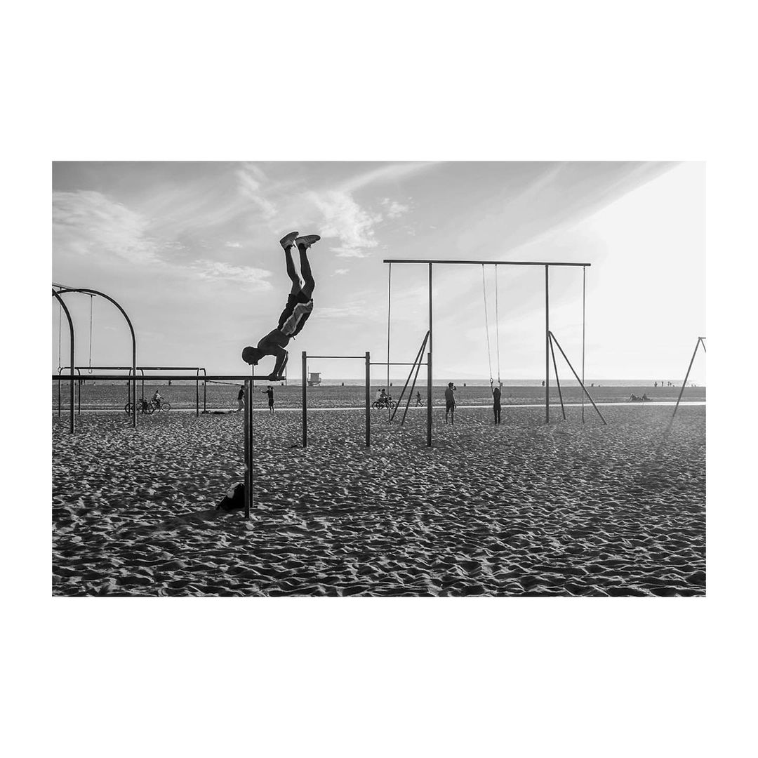 Monochrome shot of a person doing a handstand on a bar at an outdoor beach gym, with other workout structures and people in the background.