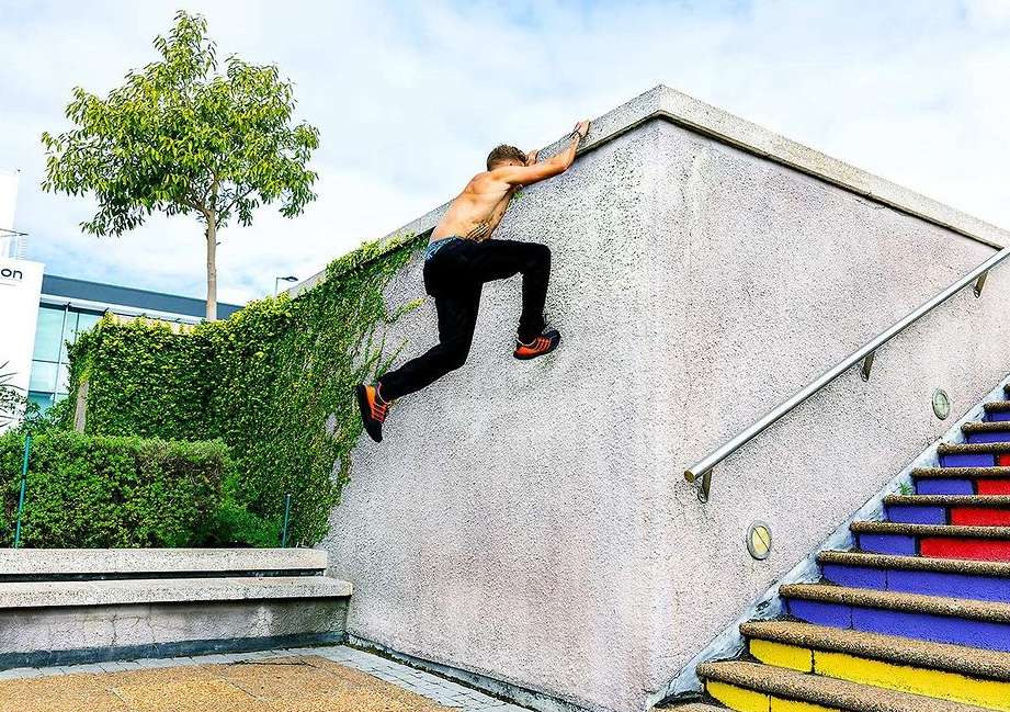 An adult male doing parkour on a concrete wall next to colorful stairs, wearing black pants and orange shoes in an urban environment.