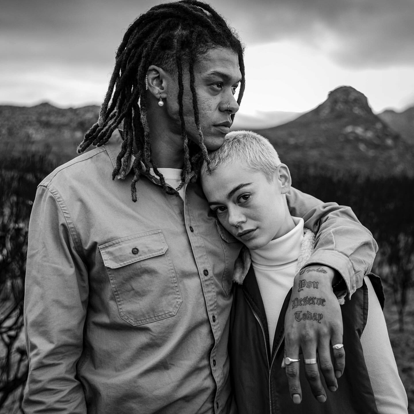 Black and white portrait of a man with dreadlocks holding a short-haired woman resting her head on his shoulder outdoors.