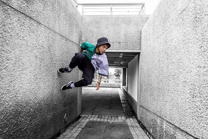 Young man mid-air parkour jump off a textured concrete wall wearing a bucket hat, green and white jersey, and black pants.