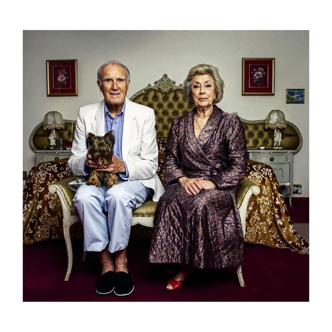 Elderly couple sits with a Yorkie on a bed in a bedroom, with the man wearing a white jacket and the woman in a quilted robe.