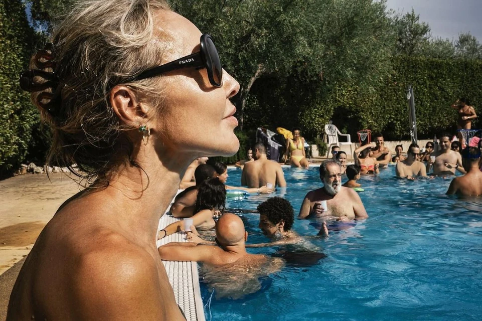 Woman in Prada sunglasses relaxes at crowded pool party. Lush greenery surrounds the outdoor summer scene with clear blue water.