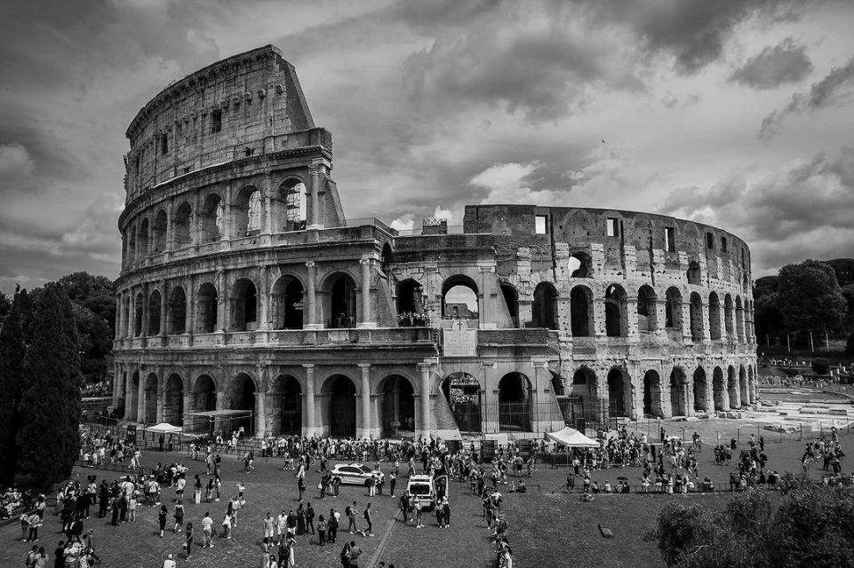 Monochrome photograph of the Colosseum in Rome, Italy, under a cloudy sky, with crowds of people on the ground.