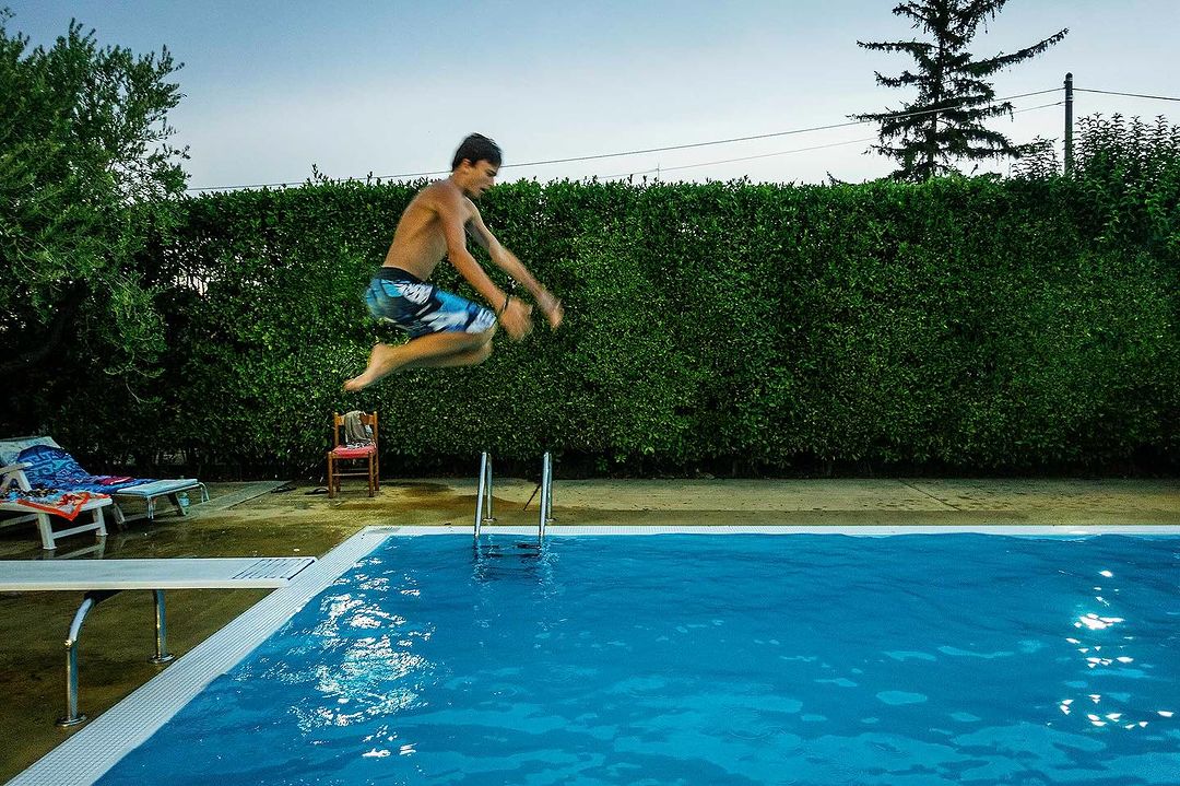 Teenage boy in mid-air jumping into a blue outdoor swimming pool. Diving board and green hedge are also visible. 