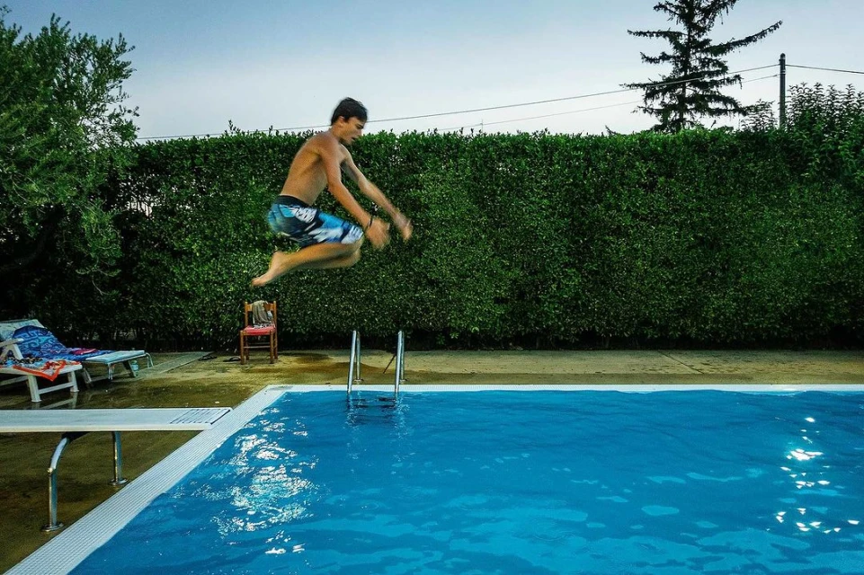 Teenage boy in mid-air jumping into a blue outdoor swimming pool. Diving board and green hedge are also visible. 
