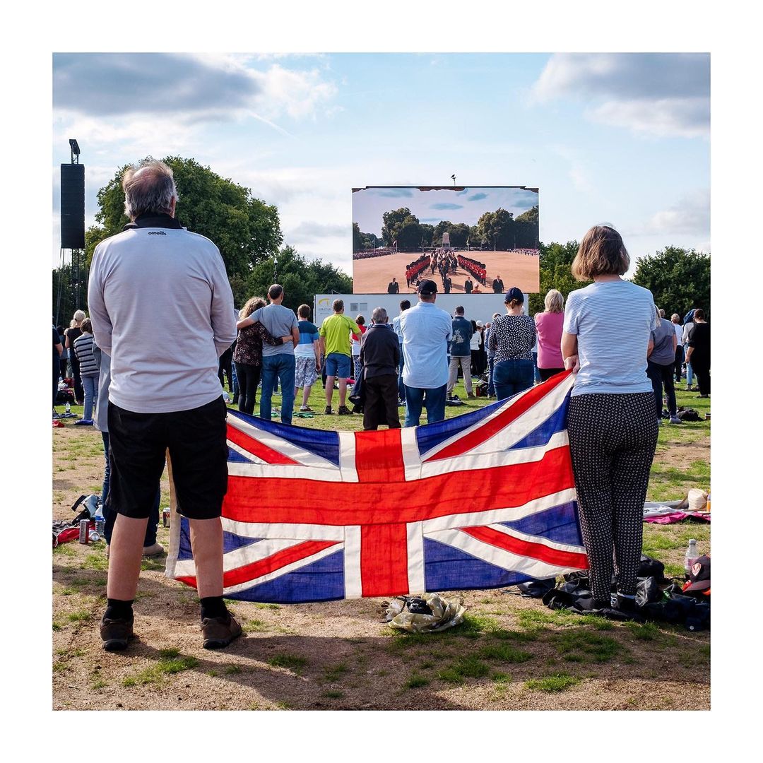 People watch a large outdoor screen displaying a royal event, with a Union Jack flag draped in front on a grassy field under sky.