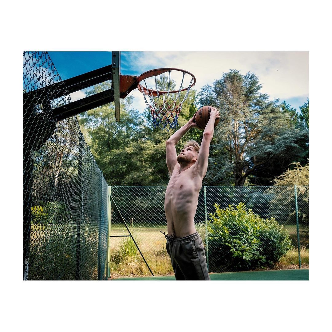 An adult male with a basketball in the air, preparing to shoot at an outdoor hoop against a backdrop of green trees and a blue sky.