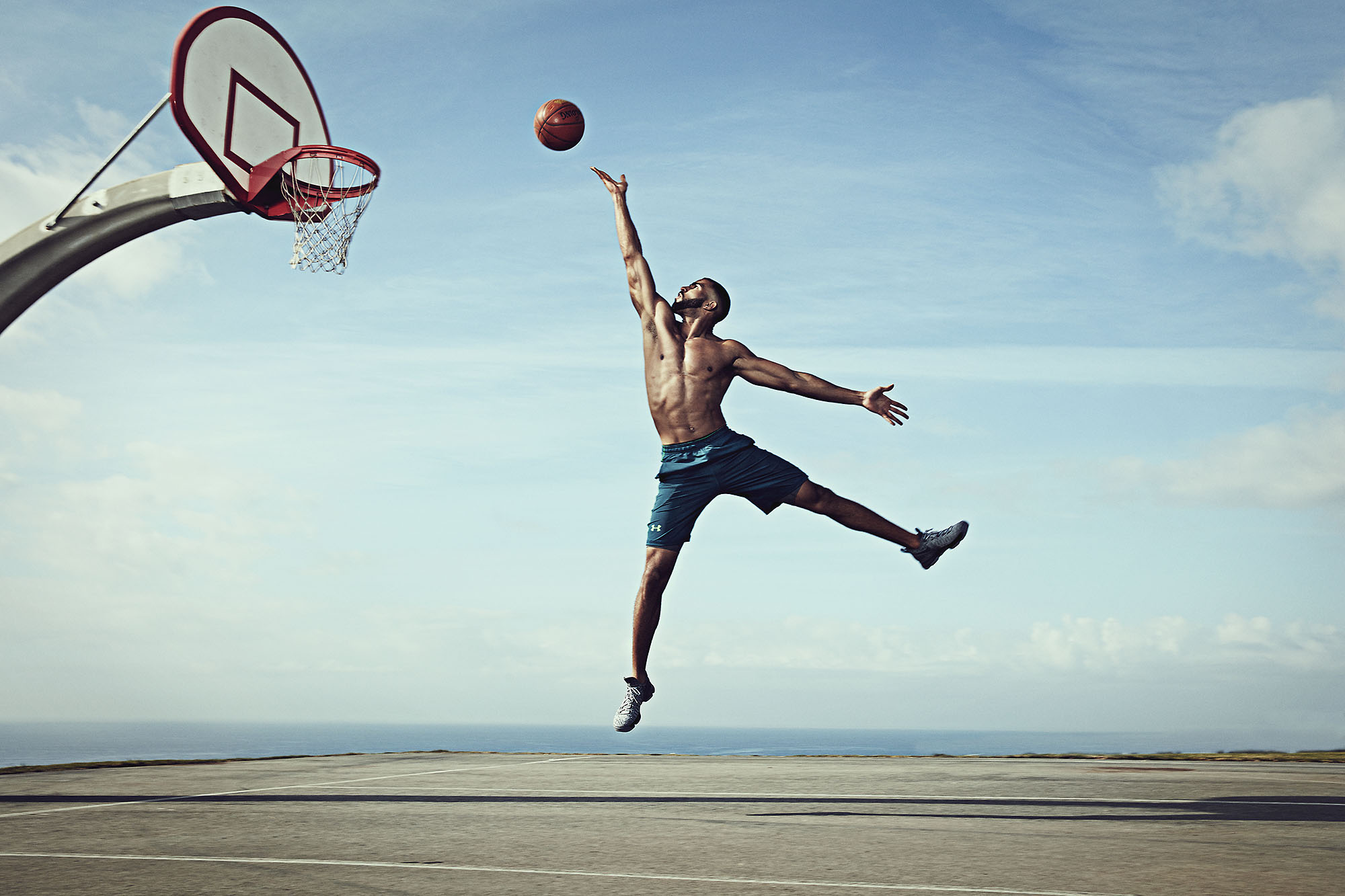 An athletic man leaps to dunk a basketball on an outdoor court, set against a bright blue sky and distant ocean backdrop, under natural light.