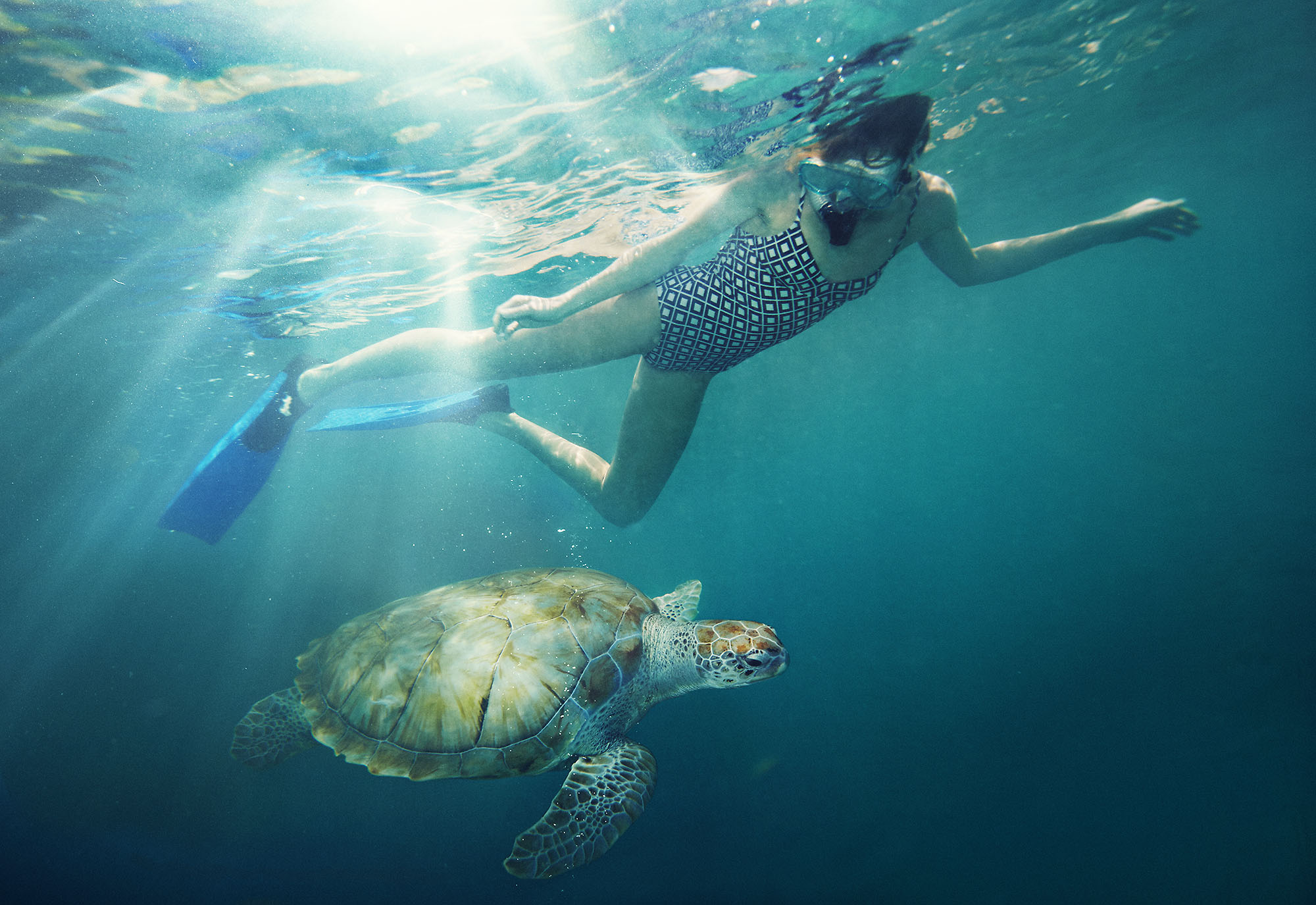 Underwater photography of an adult female snorkeling alongside a sea turtle in a tropical ocean with clear blue water and sun rays.