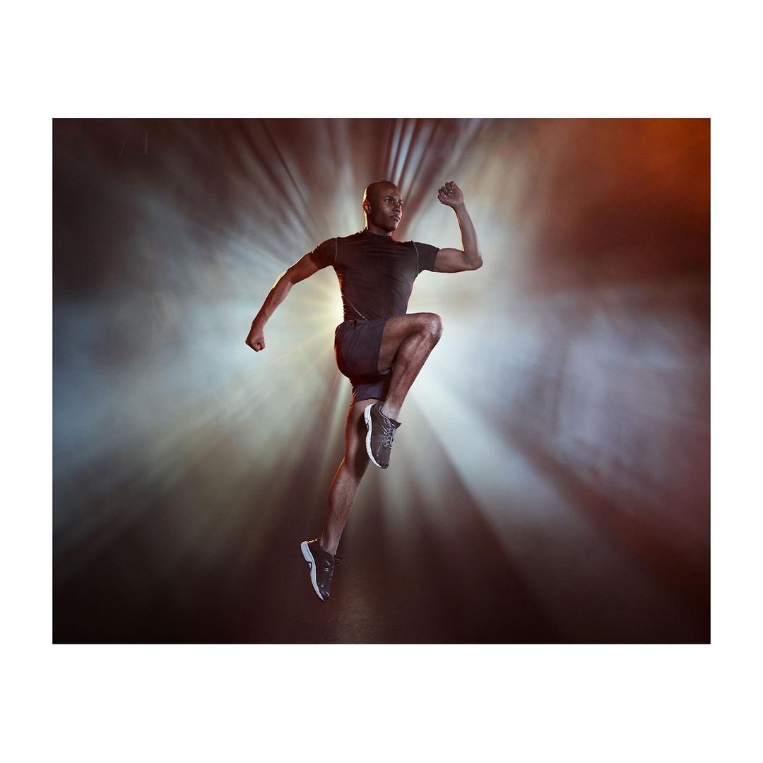 Athletic Black man in sportswear dynamically leaping against a bright, diffused spotlight background, showcasing motion and physique.