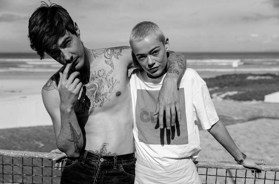 Black-and-white portrait of a shirtless man with tattoos and a woman in a t-shirt hugging near a railing with a beach background.