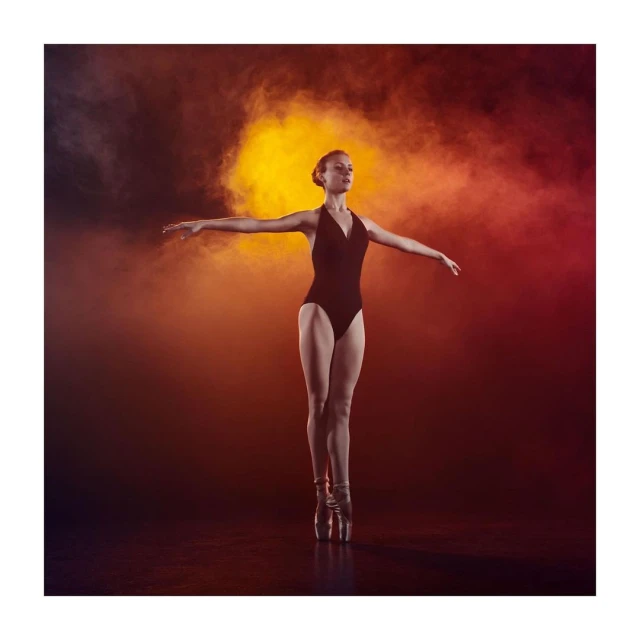 Female ballet dancer in leotard and pointe shoes poses en pointe against a backdrop with dramatic orange and black smoke.