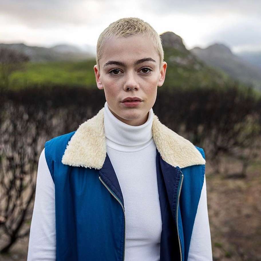 Portrait of a young adult female with buzzcut, wearing a white turtleneck and blue vest against an outdoor landscape background.