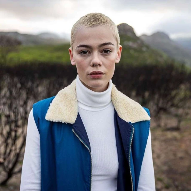 Portrait of a young adult female with buzzcut, wearing a white turtleneck and blue vest against an outdoor landscape background.