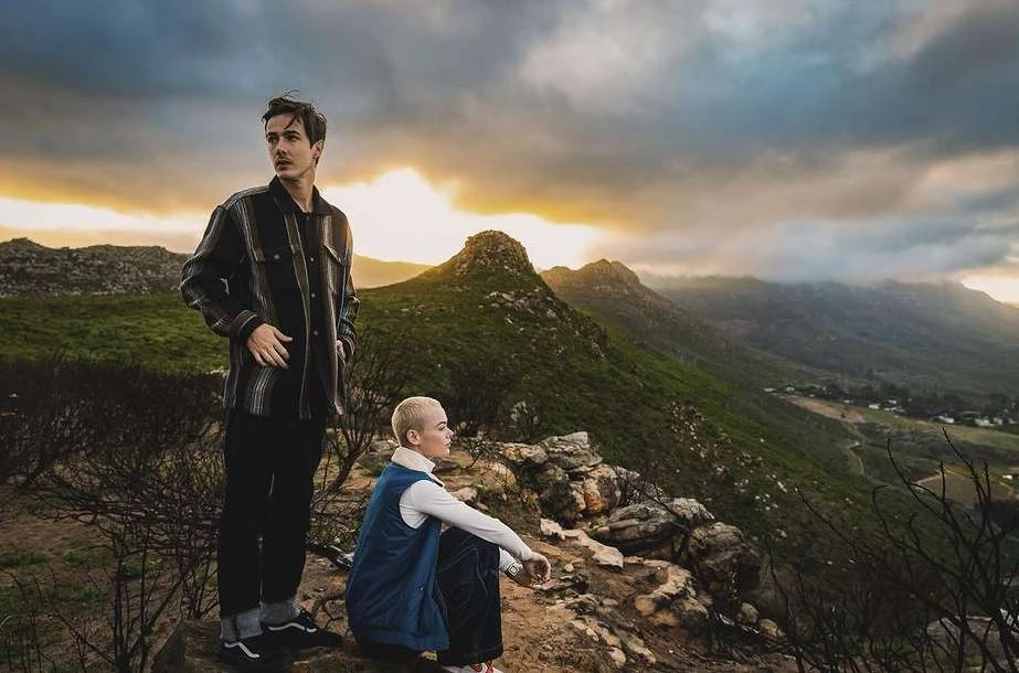 Outdoor portrait of a man standing and woman sitting in nature, capturing a mountain landscape under dramatic cloudy skies at sunset.