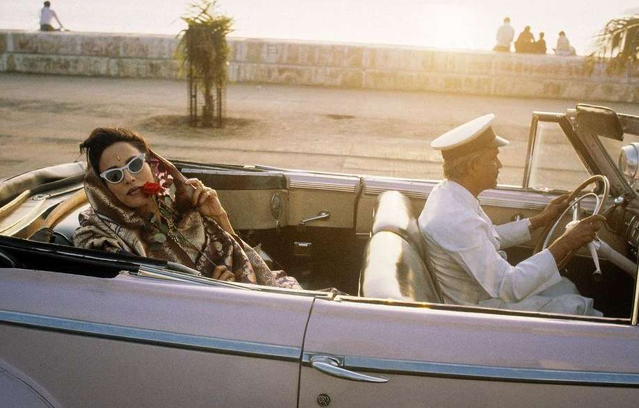 A woman and driver in an open-top car with rose, sunglasses, and white cap against a sunlit coastal background, exuding vintage style.