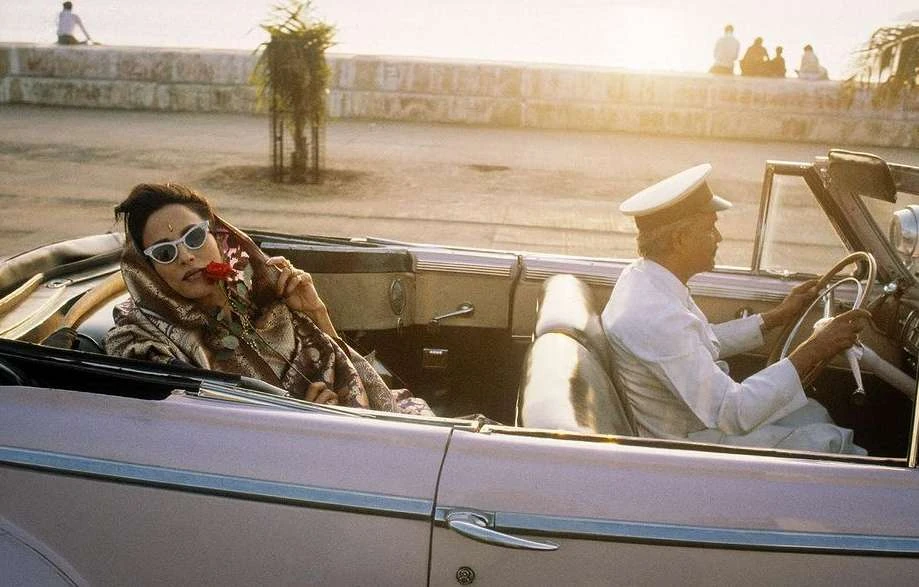 A woman and driver in an open-top car with rose, sunglasses, and white cap against a sunlit coastal background, exuding vintage style.