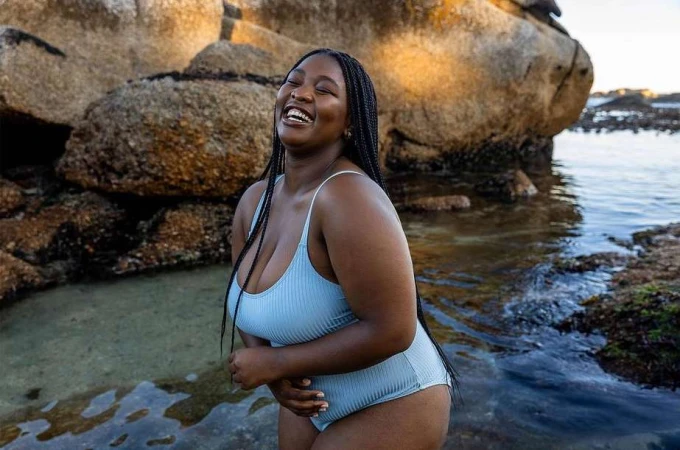 A joyful dark-skinned woman in a light blue swimsuit stands waist-deep in water near rocks on a sunny day, laughing with her eyes closed.