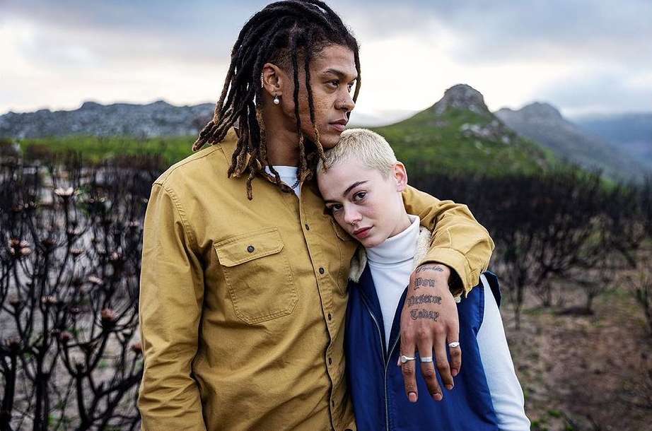 Man with dreadlocks wearing a yellow shirt and woman with short blonde hair embracing outdoors.