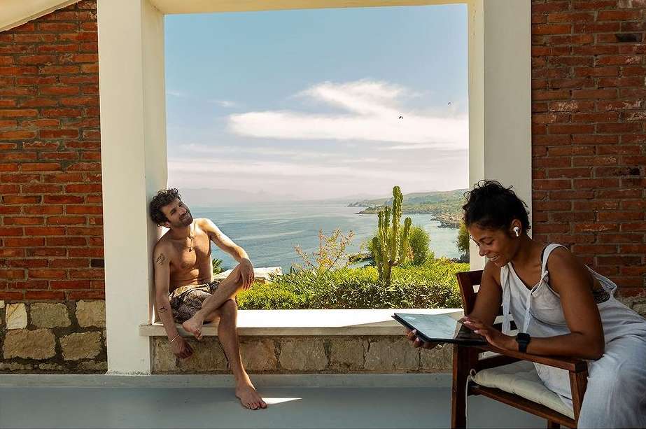 A man relaxes on a ledge with a sea view while a woman uses a tablet, both enjoying a bright, sunny day outdoors.