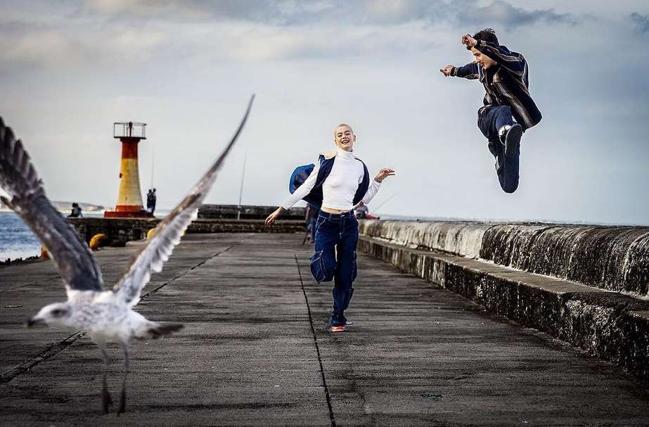 Two people outdoors on a concrete pier with a lighthouse and cloudy sky in the background and a seagull in the foreground.