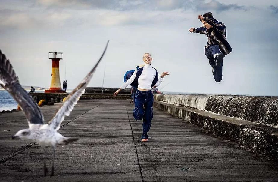 Two people outdoors on a concrete pier with a lighthouse and cloudy sky in the background and a seagull in the foreground.