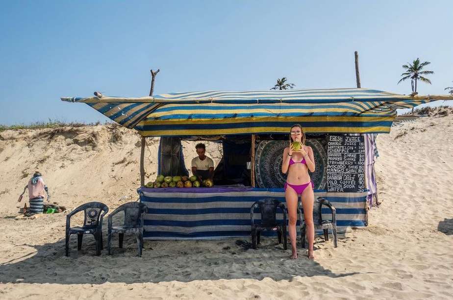 Woman in bikini drinking coconut juice at beach stand, vendor and coconuts visible, sandy beach and clear blue sky backdrop.
