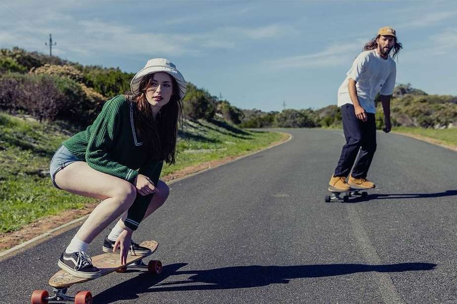 Two people longboarding down an asphalt road on a sunny day, with a grassy hill in the background under a blue sky.