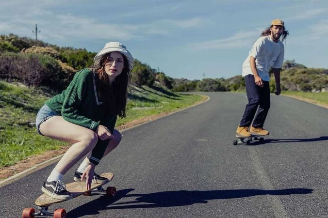 Two people longboarding down an asphalt road on a sunny day, with a grassy hill in the background under a blue sky.