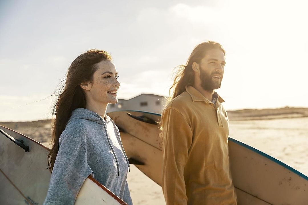 A smiling couple walks on a beach with surfboards on a sunny day. The man has long hair and a beard; the woman has long brown hair.
