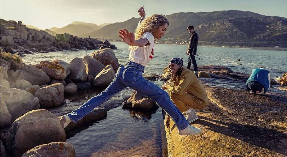 Woman jumps a stream on a rocky coast near the ocean with mountains in background. People are relaxing nearby in the sunshine.